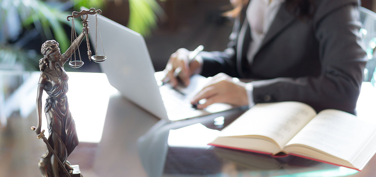 A statue of Lady Justice sits on a desk in the foreground, while in the background is a business woman working on a laptop.